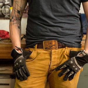 May include: A person wearing a gray t-shirt and brown pants with a belt buckle featuring a stylized animal design. The person is wearing black gloves with silver accents.