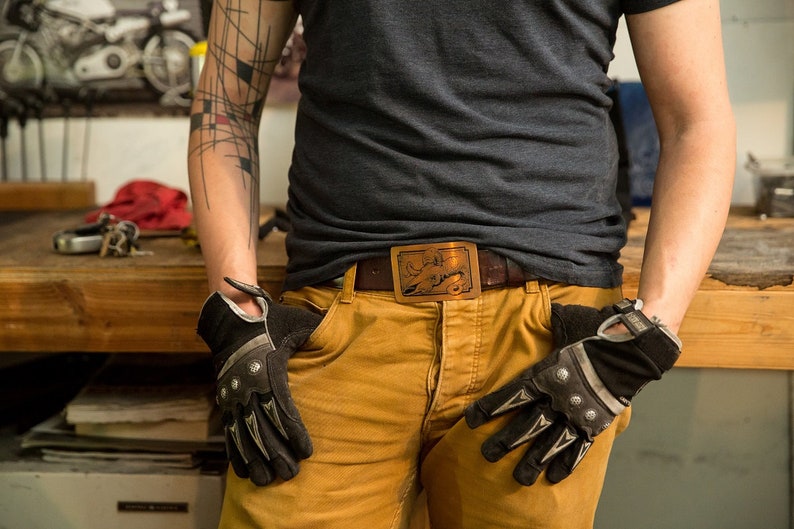 May include: A person wearing a gray t-shirt and brown pants with a belt buckle featuring a stylized animal design. The person is wearing black gloves with silver accents.