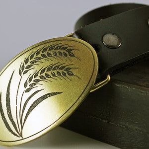 May include: A gold-colored oval belt buckle with a black wheat design. The buckle is attached to a black leather belt.