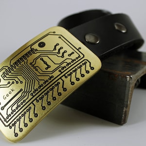May include: A gold-colored belt buckle with a black circuit board design and the word "Geek" printed on it. The buckle is attached to a black leather belt.
