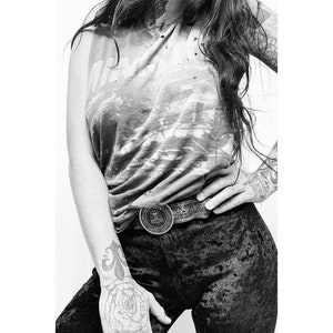 May include: A black and white photo of a person wearing a black velvet pants and a sleeveless shirt with a faded graphic print. The person is wearing a silver belt with a circular buckle.