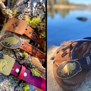 May include: Two leather belts with metal belt buckles. The buckles are oval shaped and feature a gold-toned buffalo silhouette design. One belt is brown with a dark brown leather buckle and the other is red with a gold-toned buckle.