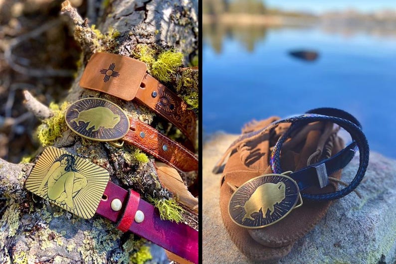 May include: Two leather belts with metal belt buckles. The buckles are oval shaped and feature a gold-toned buffalo silhouette design. One belt is brown with a dark brown leather buckle and the other is red with a gold-toned buckle.