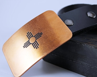 May include: A copper belt buckle with a black Zia symbol, a traditional symbol of the Pueblo people of New Mexico, on a black leather belt.