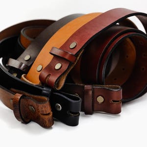 May include: Four leather belts in different shades of brown, with a buckle on each. The belts are stacked on top of each other.