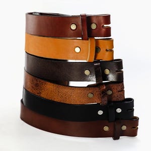 May include: A stack of six leather belts in various shades of brown, black, and tan. The belts are all made of leather and have a simple design with a single buckle.