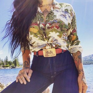 May include: A woman wearing a white and green patterned button-down shirt tied at the waist, a red belt with a gold buckle featuring a bear and the text "California Republic", and blue jeans.