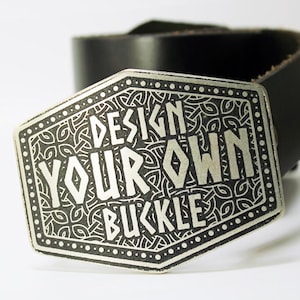 May include: A silver belt buckle with a black leather belt. The buckle has a detailed design with the text "DESIGN YOUR OWN BUCKLE" in white.