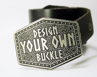 May include: A silver belt buckle with a black leather belt. The buckle has a detailed design with the text "DESIGN YOUR OWN BUCKLE" in white.