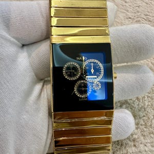 Könnte beinhalten: Eine goldfarbene Rado Jubilé Armbanduhr mit rechteckigem Gehäuse und schwarzem Zifferblatt. Das Zifferblatt enthält drei runde Hilfszifferblätter mit kleinen klaren Steinen, goldene Zeiger und das Rado-Logo. Breites, segmentiertes Goldarmband.