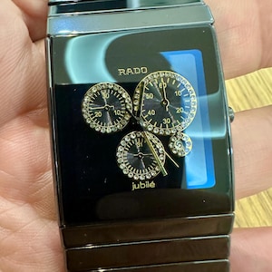 Könnte beinhalten: Eine schwarze Rado Jubilé Armbanduhr aus Keramik mit rechteckigem Zifferblatt. Das Zifferblatt zeigt drei kreisförmige Hilfszifferblätter mit kleinen klaren Steinen, goldfarbene Zeiger und das Rado-Logo. Passendes schwarzes Keramikgliederarmband.