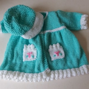 May include: A teal knitted baby dress with white trim and two white pockets with pink accents. The dress comes with a matching teal knitted hat with a white brim.