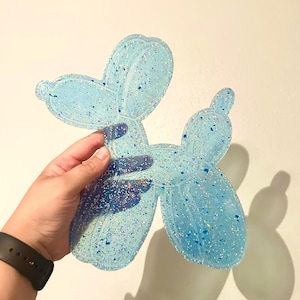 May include: A light blue, glitter-infused resin balloon dog figurine. The figurine is in the shape of a balloon animal, with a rounded head, body, and legs. The resin is translucent, allowing light to pass through, and is filled with iridescent glitter.