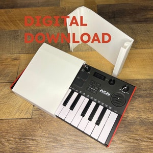 Puede incluir: Un teclado MIDI Akai Professional negro y rojo con teclas blancas está parcialmente dentro de una funda blanca. El texto "DIGITAL DOWNLOAD" está en letras rojas encima del teclado. El teclado tiene perillas y un altavoz.