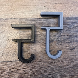 May include: Two 3D printed hooks, one black and one gray, are shown on a woodgrain surface. The hooks are shaped like a letter J with a small square at the top.