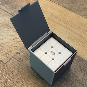 Puede incluir: Una caja de plástico gris con una tapa con bisagras, abierta para revelar un cubo de rompecabezas blanco 3x3x3 en el interior.