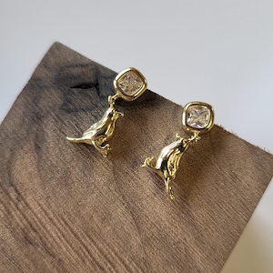Puede incluir: Un par de pendientes dorados con forma de pájaro. Cada pendiente presenta un dije con forma de pájaro que sostiene una gema cuadrada y transparente. Los pendientes se exhiben sobre una superficie de madera con un fondo claro.