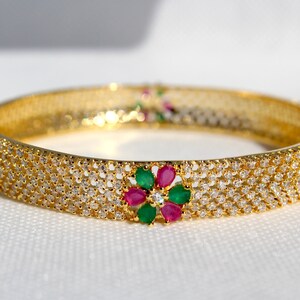 May include: A gold bangle bracelet with a row of clear cubic zirconia stones and a flower design with green and pink stones.