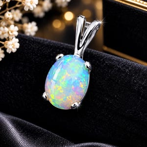 May include: A silver pendant featuring an oval-shaped, cabochon-cut opal gemstone with iridescent flashes of blue, green, and yellow. The opal is secured in a four-prong setting attached to a polished silver bail, creating a bright, reflective piece.