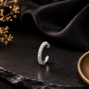 May include: A silver-toned ear cuff featuring a row of small, clear, sparkling stones set along the outer edge. The piece has a C-shaped design with a polished metallic finish, showcasing a curved band that is open at one end for placement.