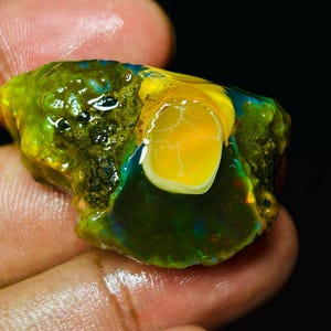 May include: A rough, green and yellow opal gemstone with a bright yellow center. The opal is iridescent and has a rough, natural texture.