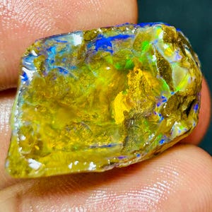 May include: A rough opal gemstone displaying a spectrum of colours. The stone has a yellow and gold base, with flashes of blue and green. The opal is held in a hand, highlighting its natural, unpolished form.