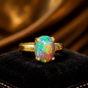 Op de afbeelding: Een gouden ring met een grote, ovaalvormige cabochon opaal edelsteen in een zetting met vier pootjes. De opaal vertoont een levendig kleurenspel met flitsen van groen, rood en blauw over het gehele oppervlak, gecombineerd met een gouden band.