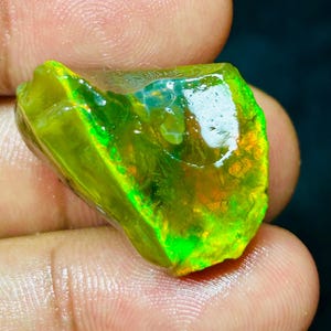 Könnte beinhalten: Ein lebendiger, roher Opal-Edelstein mit einer Mischung aus grünen, gelben und orangen Farbtönen. Der Edelstein wird in einer Hand gehalten und zeigt seine natürliche, raue Textur und das innere Farbspiel. Die Oberfläche des Opals reflektiert das Licht.