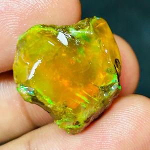 Op de afbeelding: Een ruwe, ongeslepen opaal edelsteen met een levendige kleurenpracht. De steen heeft een goudgele basis met flitsen van groen en oranje. De opaal wordt in een hand gehouden, waardoor de natuurlijke, onregelmatige vorm en unieke patronen worden getoond.