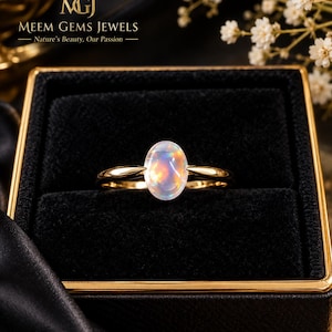 May include: A gold ring featuring a central oval-shaped cabochon opal with iridescent flashes of colour. The ring is displayed inside a black velvet-lined box. The text MGJ MEEM GEMS JEWELS Nature's Beauty, Our Passion is visible at the top.