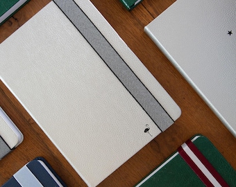 May include: Six different notebooks with various colors and designs. Each notebook has a ribbon bookmark and a unique design on the cover. The notebooks are arranged on a wooden surface.