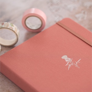 May include: A coral coloured notebook with a silver embossed thistle design. The notebook has a brown leather strap closure.