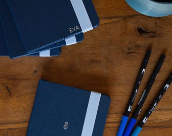 May include: Three navy blue notebooks with white elastic bands and personalized lettering. The top notebook says "EVA" and the bottom notebook has a pineapple design. Three blue pens are on a wooden surface.