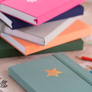 May include: A stack of five notebooks in various colors, including pink, blue, gray, orange, and green. The top notebook has a gold star on the cover. The notebooks are on a wooden surface.