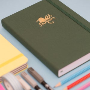 Puede incluir: Un diario verde oscuro con un diseño de pulpo dorado en la portada. El diario tiene un cierre elástico blanco y un borde de página blanco. El diario está sobre una superficie azul claro con otros artículos de papelería.