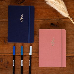 Puede incluir: Dos cuadernos con un diseño de clave de sol dorada. Un cuaderno es azul marino y el otro es rosa. Tres bolígrafos negros con tinta azul, verde azulado y blanca están sobre una superficie de madera.