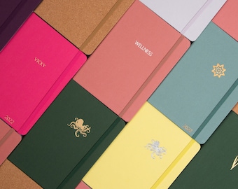 May include: A collection of colorful notebooks with various designs and text, including "Wellness", "Vicky", and "2022".