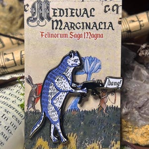 Pode incluir: Um pin de esmalte "Medieval Marginalia" com um gato azul e branco segurando uma metralhadora com um balão "bang!". A ilustração medieval inclui um coelho e outras figuras.