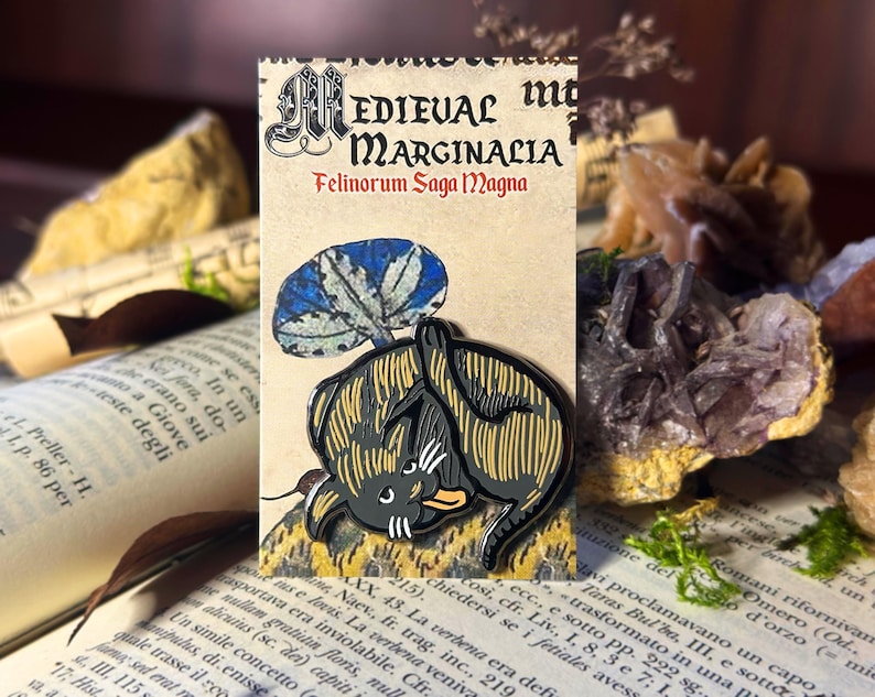 May include: A black and gold enamel pin depicting a cat with a blue and white mushroom on its back. The pin is on a card with the text "Medieval Marginalia Felinorum Saga Magna".
