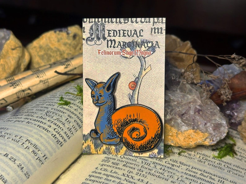 May include: A blue and orange enamel pin depicting a cat with a snail shell on its back. The pin is set against a background of a medieval-style illustration with the text "Medieval Marginalia Felinorum Saga Magna".