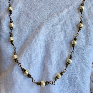 May include: A long necklace with faux pearls and antique brass-coloured wire. The necklace has a delicate design with evenly spaced, round, off-white pearls. The wire is intricately wrapped around the pearls and connecting elements.