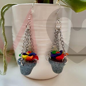 Puede incluir: Pendientes hechos a mano con macetas grises brillantes en miniatura y flores coloridas. Los pendientes cuelgan de cadenas y ganchos plateados, creando un accesorio único. Las flores son una mezcla de rojo, amarillo, azul y verde.