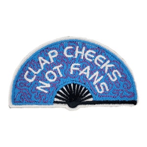 May include: A blue and white embroidered patch shaped like a fan with the text "Clap Cheeks Not Fans" in white.