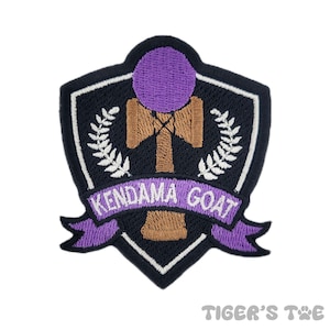 Puede incluir: Parche bordado morado y marrón con un kendama, un juguete tradicional japonés, y el texto "Kendama Goat" en una pancarta blanca.