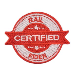May include: Embroidered patch with the text "RAIL CERTIFIED RIDER". The patch is circular with a red and white design. The words are in white, set against a red banner. The patch has a white border and star designs above and below the banner.
