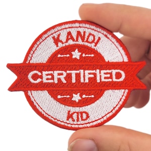 May include: Embroidered patch with a red and white design. The patch features the words "KANDI CERTIFIED KID" in white lettering. The patch is circular with a red banner across the centre. The patch is held by a hand.