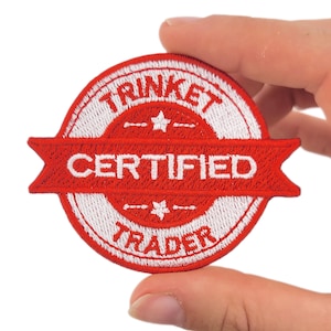 May include: A circular, red and white embroidered patch with the text "TRINKET CERTIFIED TRADER". The patch features a banner design with the word "CERTIFIED" in white. The patch is held by a hand.
