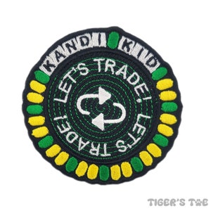 May include: Circular embroidered patch with the text "KANDI KID" in white, and "LET'S TRADE!" in white. The center features a white arrow symbol. The patch has a black background with yellow and green accents.