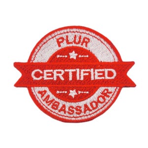 May include: A circular red and white embroidered patch with the words "PLUR CERTIFIED AMBASSADOR". The patch has a red banner across the center with white text and a white border.