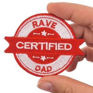May include: A red and white embroidered patch with the words "RAVE CERTIFIED DAD." The patch has a circular design with a banner across the centre. The text is in white, and the patch is held by a hand.